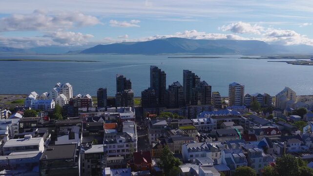 Aerial view of Reykjavik harbor, Faxafloi Bay, and Mount Esja. A boat moves across the sea, with low rise houses and dark waterfront towers in clear daylight.