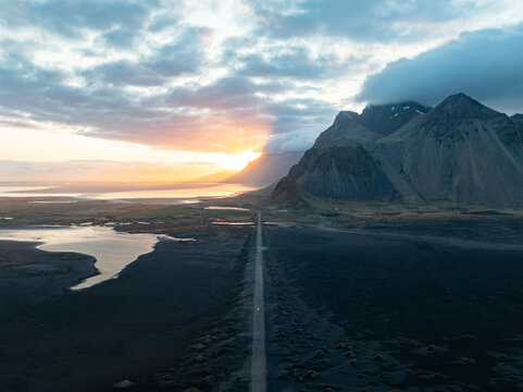 Aerial view of Stokksnes and the Vestrahorn mountain range at sunset with a long road stretching across the dark volcanic landscape in Iceland.