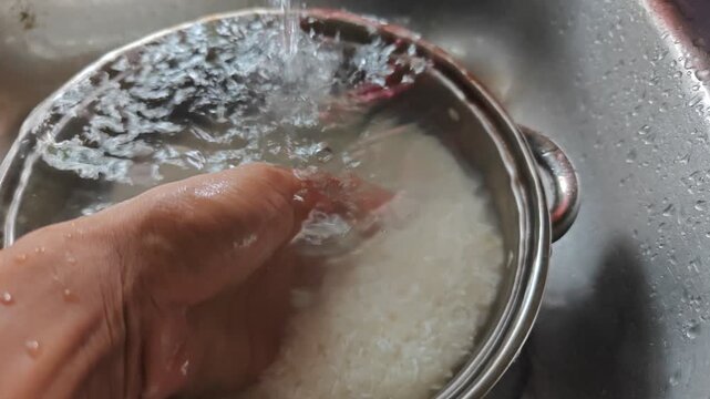 Washing raw white rice grains in a metal bowl with water splashing over a hand