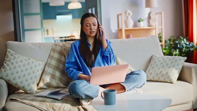 Asian freelancer phone call browsing laptop sofa. Remote worker multitasking home.