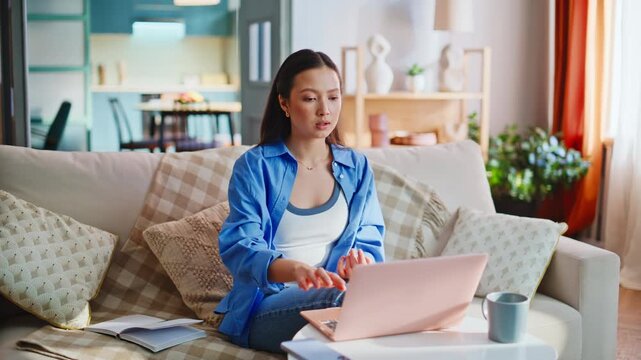Emotional asian girl video call home sofa, gesturing talking on pink laptop