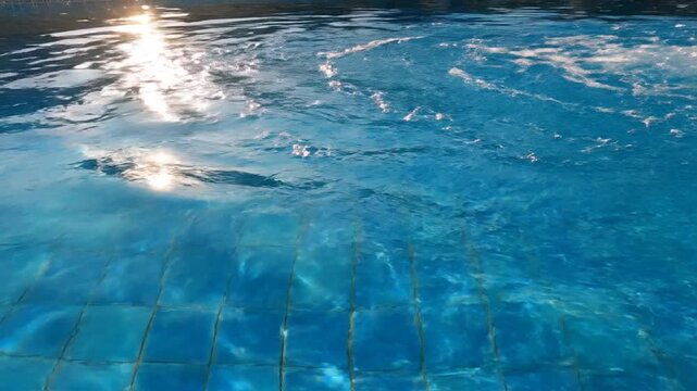Sunlight reflects on rippled swimming pool water surface with blue tiles