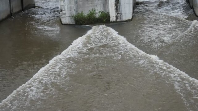 Mud flows beneath concrete bridge supports. Ripples crash against weathered pillars. Floodwaters surge with muddy force. Green shoots cling stubbornly to concrete