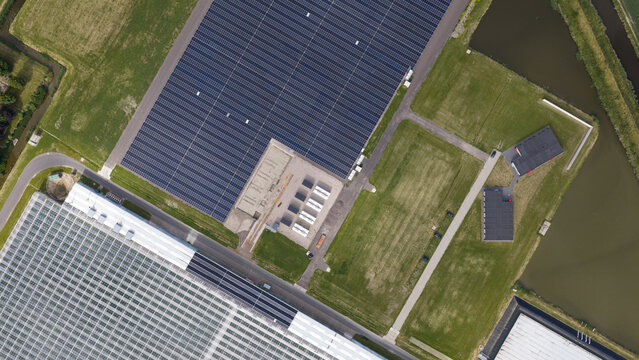 Aerial view of a large solar farm installation adjacent to industrial greenhouses, surrounded by green fields and water canals in Middenmeer, North Holland, Netherlands.