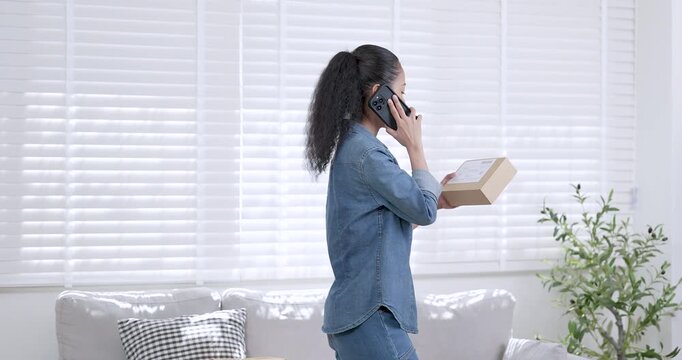 Asian woman small business owner talking on mobile phone while holding online shopping parcel box and reviewing order information showing e-commerce customer communication and online selling workflow