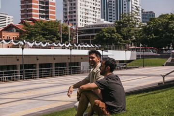 Fototapeta premium Two smiling men relaxing on urban grass, having a friendly conversation in a city park with buildings in the background