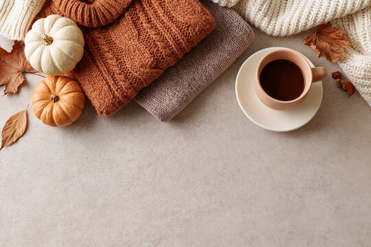 Cozy knitted sweaters rest beside tiny pumpkins. Cup of tea sits on a plain saucer. Dry leaves frame the warm autumn scene. Soft tones evoke calm, comfort, and seasonal peace. Perfect for lifestyle
