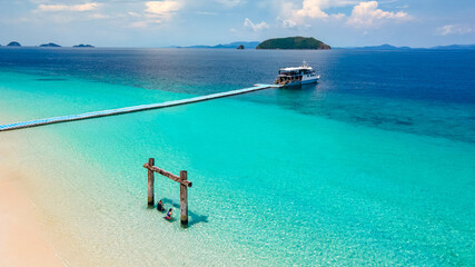 A couple tourists sitting on wooden swing on Madam beach of Nyaung Oo Phee Island in Myanmar. Tropical view of turquoise clear water and white sand with tour boat anchoring on a floating dock behind. © zephyr_p
