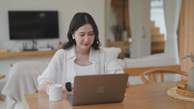 Asian woman wearing earbuds working on laptop and talking during an online learning or consulting session at home, working remotely with coffee at a wooden table in a modern room