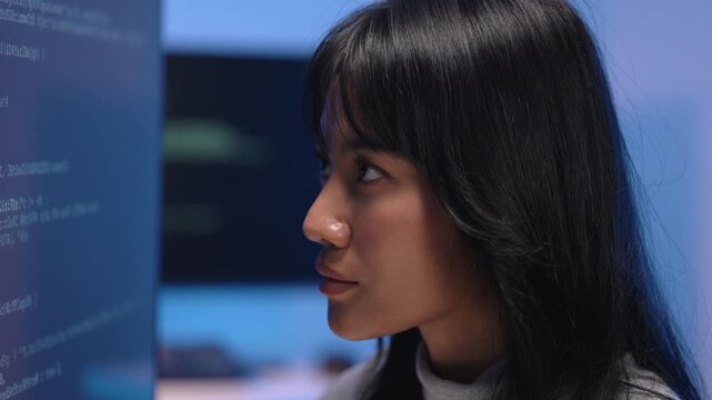 Side profile of Asian woman reading data and learning to code on computer screens in dark tech room, studying programming and analyzing information on monitor at night