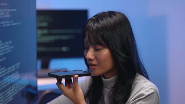 Asian woman programmer speaking into smartphone using voice AI assistant while coding, testing speech recognition technology in tech office with code on screens