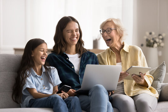 Joyful multiple generation family women using wireless devices, hold digital tablet, laptop and smartphones, laughing on joke, having fun, spend weekend on internet seated on comfortable couch at home