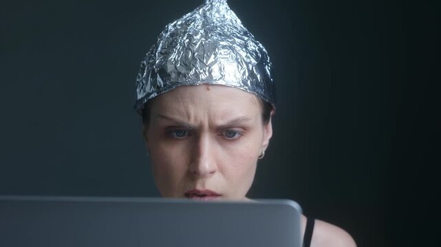 Anxious woman wearing tin foil hat staring intensely at her laptop screen, absorbed by conspiracy theories, fake news, disinformation while protecting herself from mind control. Paranoid female.