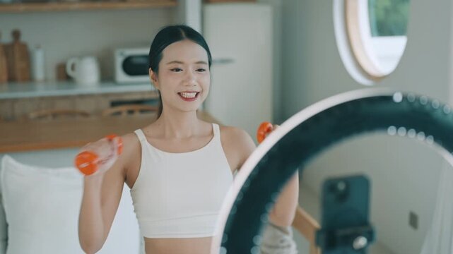 Young Asian woman smiling and talking to smartphone camera with ring light, recording vlog or live streaming wellness content at home.