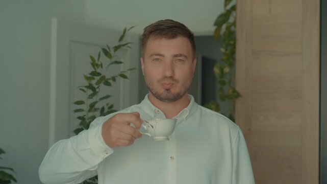 A man in a white shirt looks at the camera and drinks a drink from a tea cup. Coffee time at home.