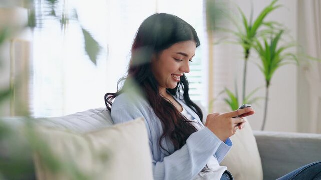 Happy young asian woman relax on comfortable couch at home texting messaging on smartphone, smiling girl use cellphone, chatting online message, shopping online from home