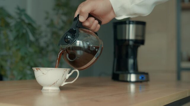 A hand pours coffee from a pot into a cup, there is a coffee maker in the background, steam is coming out of the coffee cup.