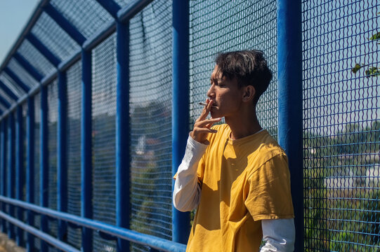 Asian young man smoking a cigarette standing by a blue bridge wire fence under bright sunlight. Concept of stress, contemplation, modern youth lifestyle, or bad habit.