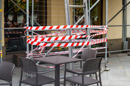 Close-up shot of an outdoor cafe seating area on the sidewalk, blocked off by metal scaffolding and construction netting wrapped in multiple layers of red and white hazard warning tape, against a beig