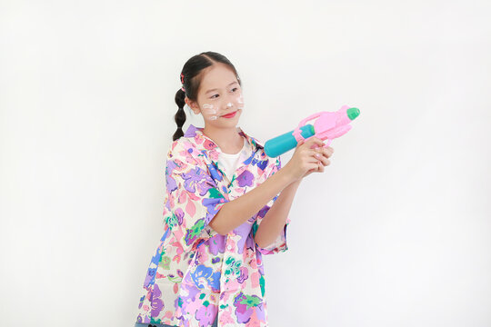 Cheerful young Thai girl with white powder on her cheeks enjoys the Songkran festival, aiming her water gun and smiling while wearing vibrant floral summer shirt.