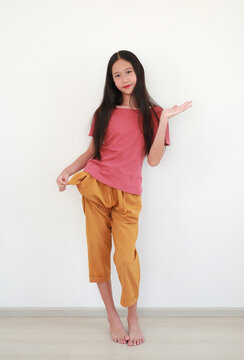Smiling young Asian girl with long dark hair in casual clothes poses playfully while holding empty pocket of her pants against white wall.