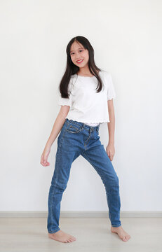 Cheerful young Asian girl striking playful and confident pose while standing against white wall, dressed in casual white short-sleeved top and blue denim jeans, smiling brightly.