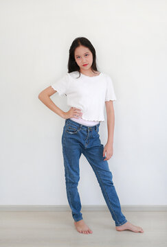 Portrait confident young Asian girl in white wavy-edged t-shirt and blue jeans, posing barefoot with her hand on her hip against white wall background.
