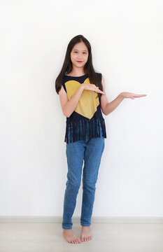 Portrait young Asian girl wearing navy blue top with a yellow heart and fringe detail paired with blue denim jeans, smiling and gesturing with her hands while posing against white wall.