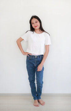 A smiling young Asian girl in casual white t-shirt and blue jeans posing confidently against white wall in the room.