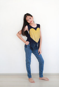 Stylish young Asian girl posing confidently against white wall, wearing a navy blue fringe top with a large yellow heart and blue jeans.