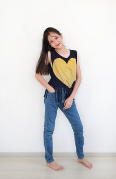 Cheerful young Asian girl posing confidently against white wall with her hand in her jeans pocket and tilting her head with a sweet smile.