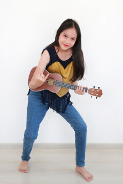 Playful Asian girl stands against white wall, strumming a ukulele like a guitar and confidently making the "rock on" hand sign, fully engaged in a energetic musical performance.