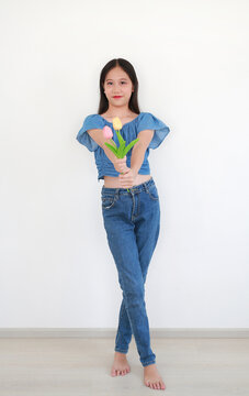 Portrait of smiling young Asian girl in stylish denim outfit posing gracefully while holding small bouquet of colorful tulips against white background.