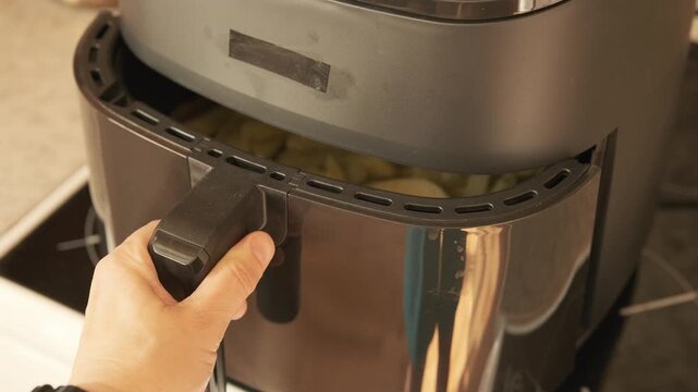 hand puts basket with raw potato fries into air fryer and presses start button for easy cooking. air fryer simplifies home cooking process. convenient air fryer makes healthy french fries