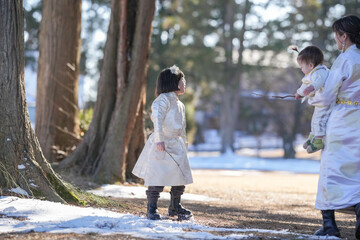 Obraz premium Soft sunlight bathes a snowy forest path. Woman cradles a baby, both dressed in white. Child stands beside them, gazing upward. Trees tower behind, with moss clinging to trunks. Snow dusts the ground