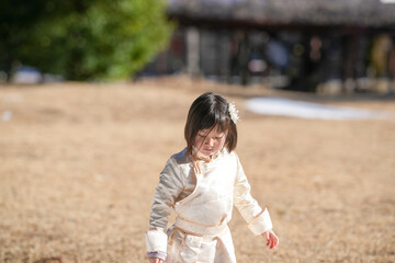 Fototapeta premium Little girl walks barefoot through golden grass. Her eyes are downcast, showing quiet focus. Sunlight bathes her in soft, warm glow. Background holds blurred trees and wooden structure