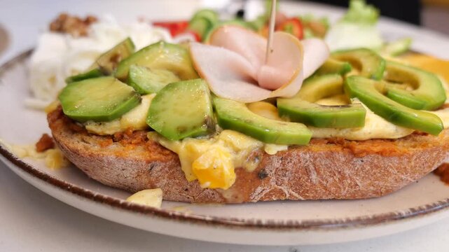 open faced sandwich featuring toasted bread topped with avocado, melted cheese, and ham