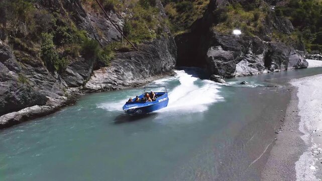 A jet boat with passengers navigates a narrow river gorge, speeding past rocky cliffs and creating a powerful wake.