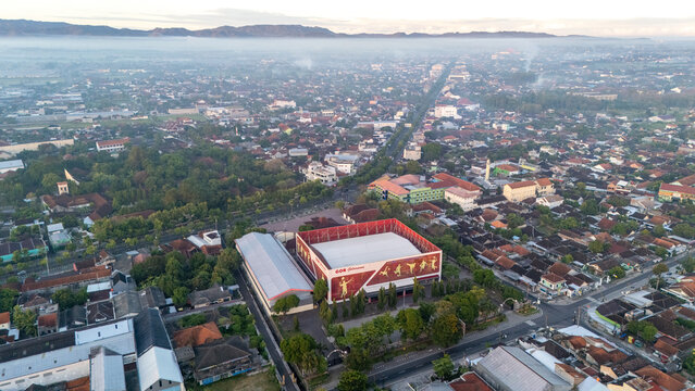 Kecamatan Klaten Utara, Indonesia - 02 July 2026: Aerial view of GOR Gelarsena Klaten sports complex surrounded by dense urban residential areas, trees, and distant misty mountains.