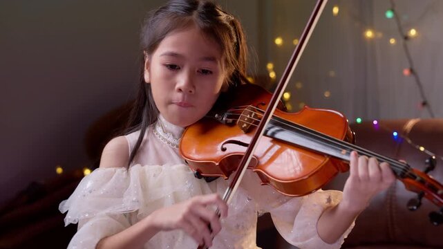 Asian girl child performing violin indoors, symbolizing education, training, discipline, and childhood in cheerful environment with warm lighting, decorative bokeh background, positive atmosphere