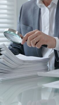 Professional person meticulously analyzing a large stack of official paperwork using a magnifying glass