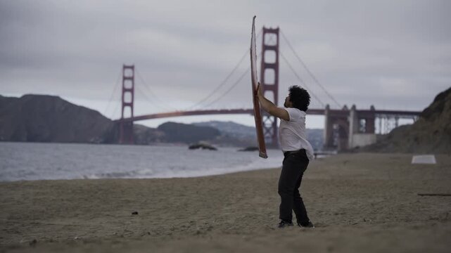 Man Spinning Large Painted Board On Beach By Golden Gate Bridge Against Strong Wind - Wide, Side Shot