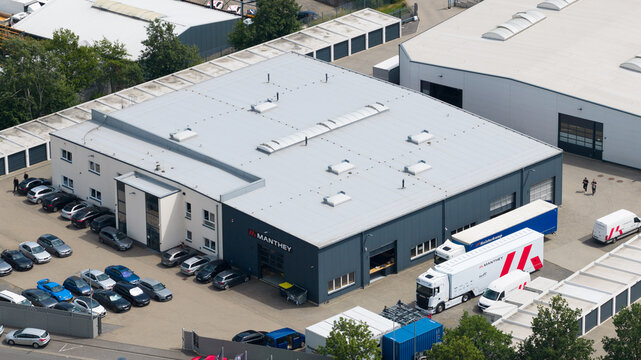 Meuspath, Germany - 23 June 2026: Aerial view of the Manthey Racing GmbH facility featuring the main industrial building, parked cars, and transport trucks in the parking area.