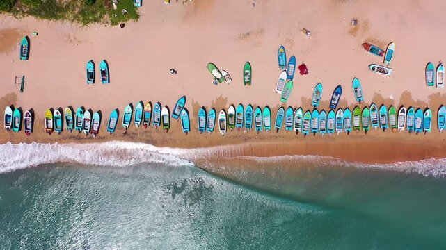 Aerial view of colorful fishing boats lined up along a sandy beach with waves from the Indian Ocean at Nilaveli, Sri Lanka. 