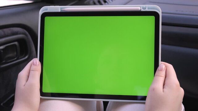 Person holds tablet with green screen inside car. Hands steady, focused on device. Ideal for app, navigation, or digital content promotion. View shows car interior details. Perfect for travel, tech