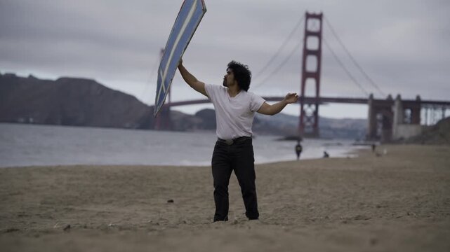 Man Swinging Painted Board On Beach In San Francisco Near Golden Gate Bridge On Windy, Stormy Day - Wide Shot