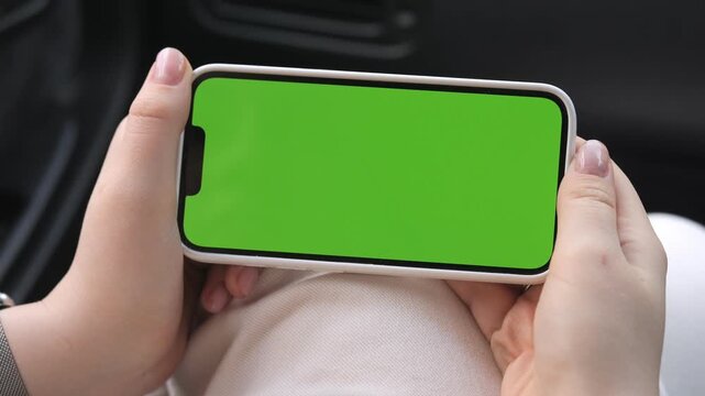 Hands hold phone with green screen inside car. Person focused on device, calm and relaxed. Ideal for apps, car tech, or digital content mockups. Green screen allows easy customization