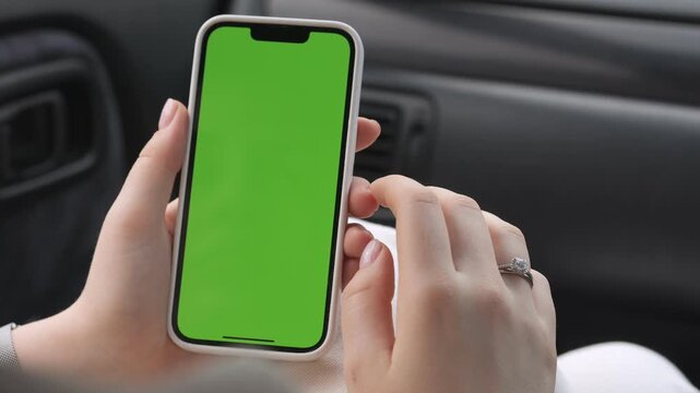Hands hold smartphone with green screen inside car. Fingers tap screen while seated. Ring visible on left hand. Green screen ready for overlay. Useful for advertising, apps, or travel content
