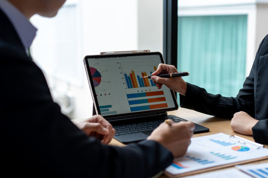 Business team reviewing sales charts on a tablet during a meeting at the office, with reports on the desk and a hand pointing at data on the screen.