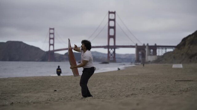 Man Dancing And Swinging Large Painted Board on Beach Near Golden Gate Bridge On Windy, Overcast Day